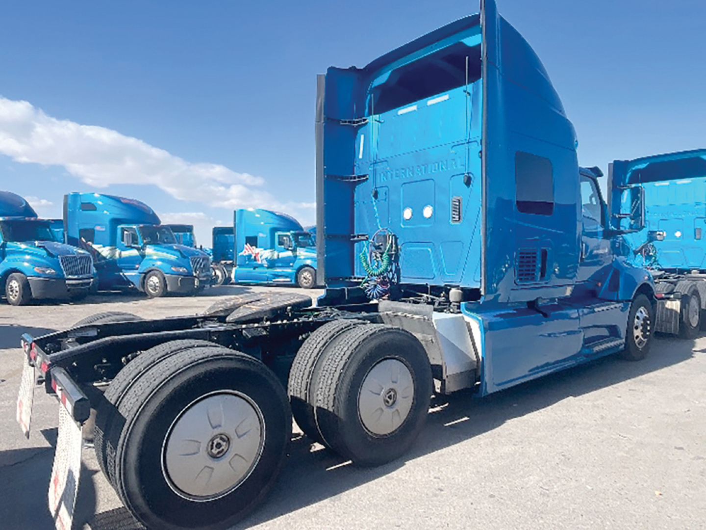 Blue semi truck tractor cab and chassis in a lot with other blue trucks; side of cab shows branding and fuel lines, rear wheels visible.