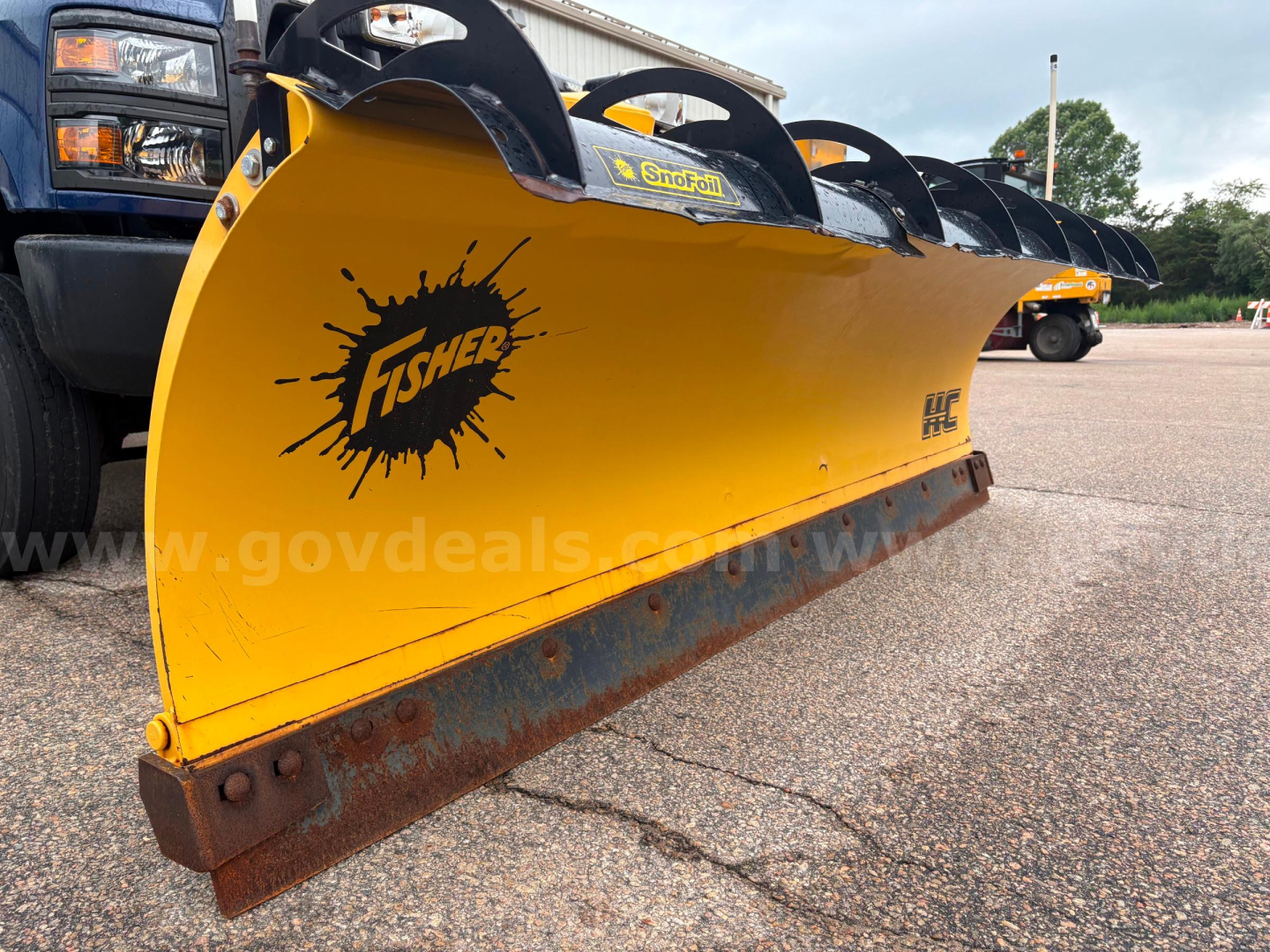 Snow plow blade attached to a dump truck with Fisher logo and yellow blade, black mounting brackets, and rusted cutting edge.