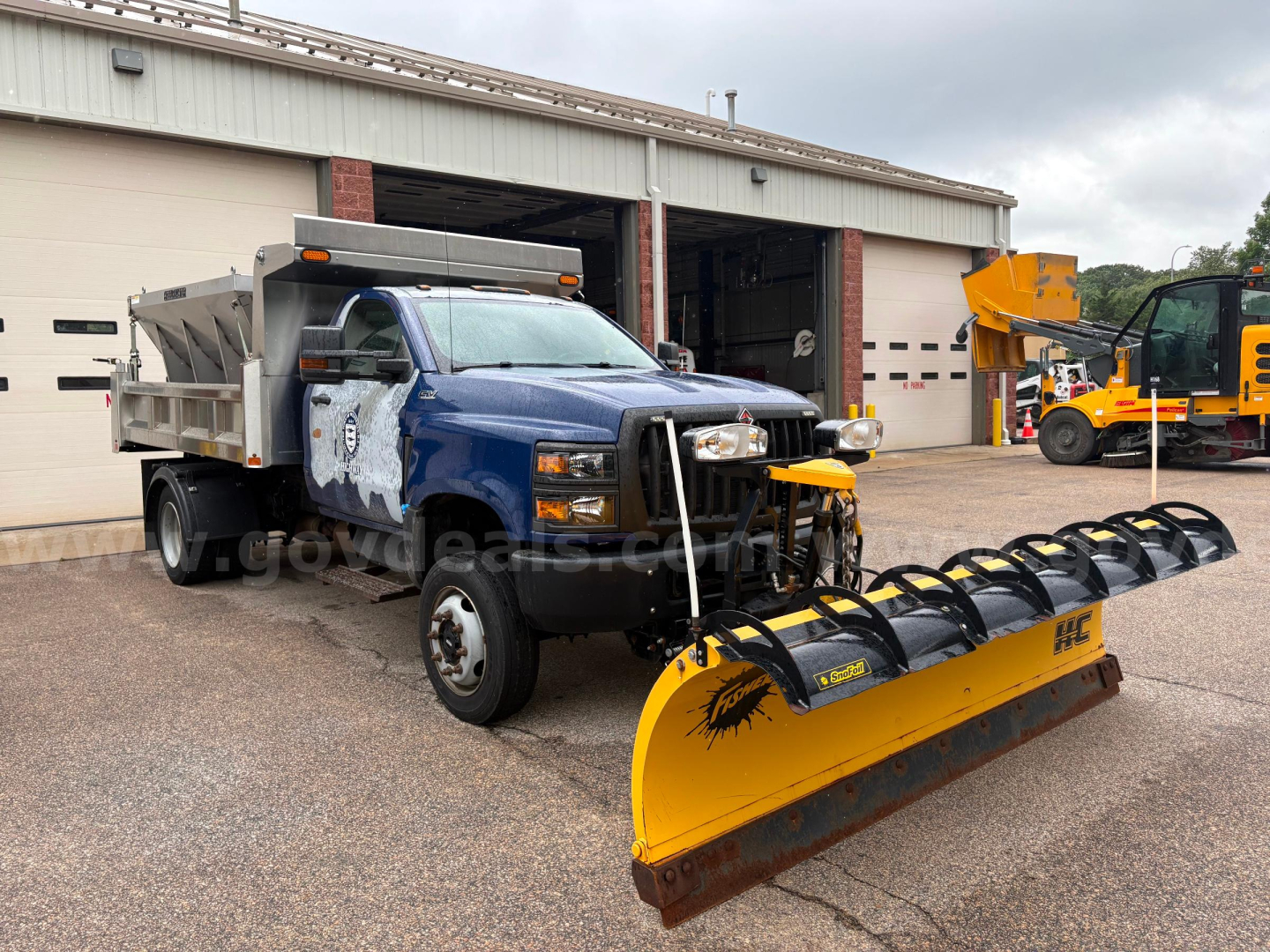 Dump truck with blue cab and yellow snow plow attachment at front, International CV515, visible in service bay.