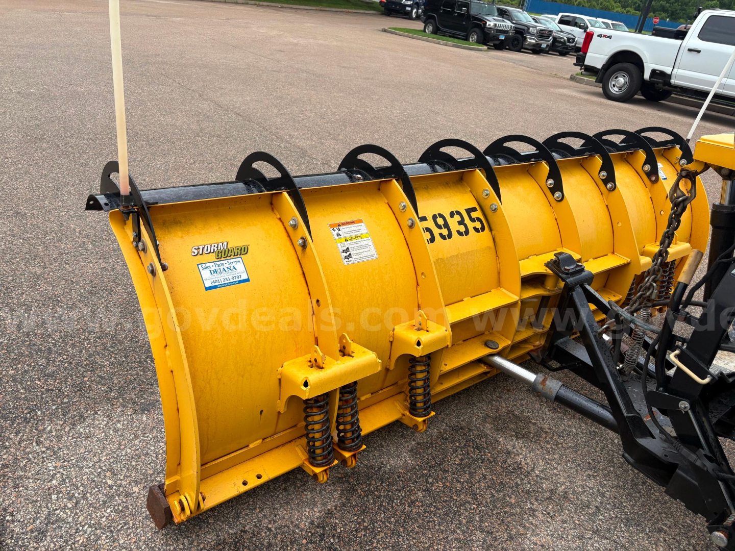 Front side of a yellow snow plow attachment with visible bolts and springs; brand label and numbers on the equipment.