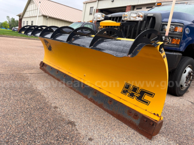 Heavy yellow snow plow attachment on front of blue truck with black blade lips and HC logo. Snow plow blade in foreground.