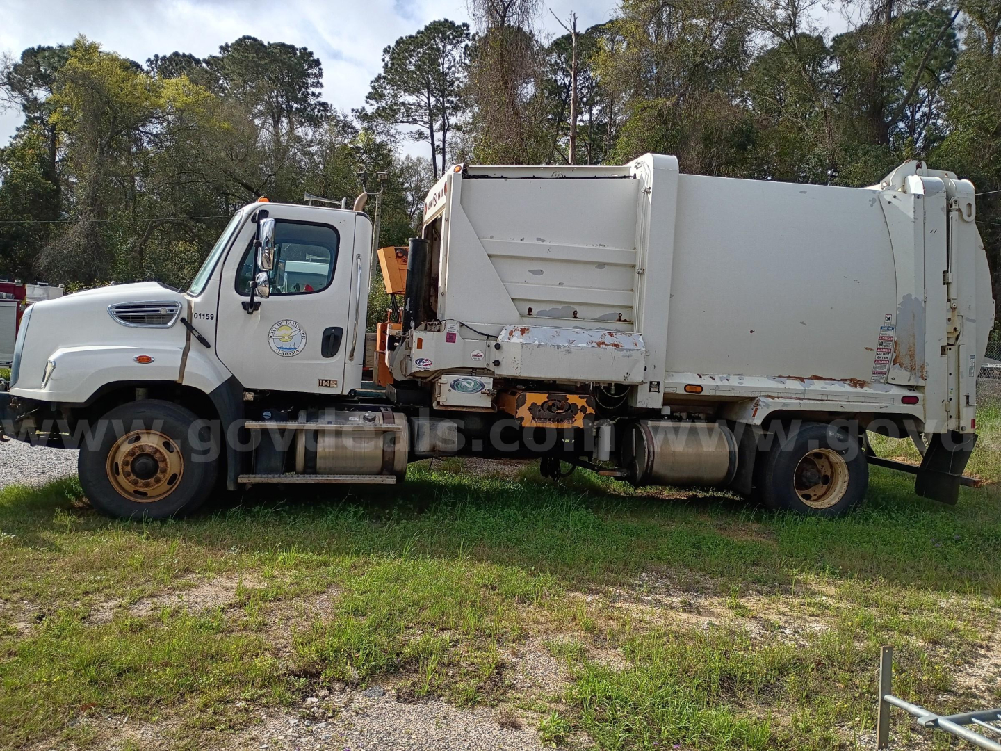 Truck side view showing 2016 Freightliner 114SD garbage truck with white cab and large rear dump body, rust spots on edges, parked on grass.