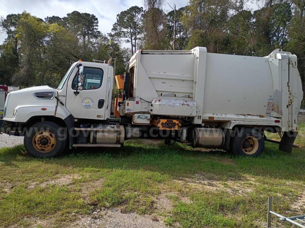 Truck side view showing 2016 Freightliner 114SD garbage truck with white cab and large rear dump body, rust spots on edges, parked on grass.