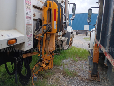 Freightliner 114SD chassis with hydraulic crane arm visible between trucks; orange/yellow crane components and control hoses; rearview mirrors and blue building in background.