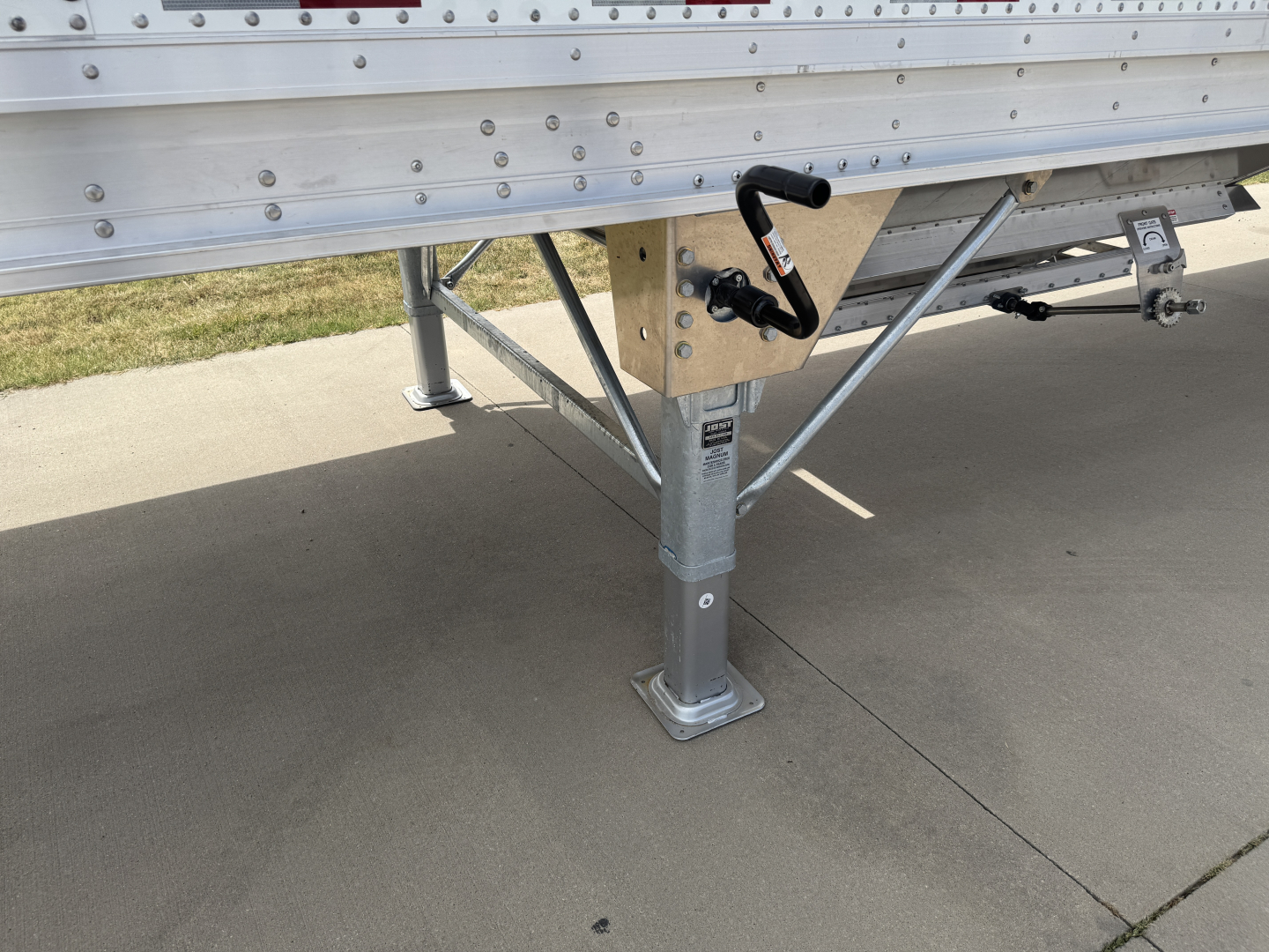 Semi-trailer hopper leg and undercarriage showing corrugated sidewall, support frame, and landing gear foot with a crank handle visible.