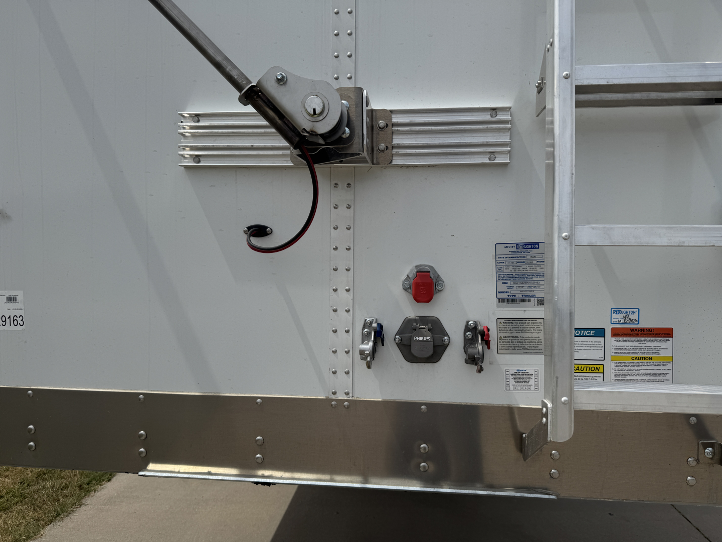 Trailer rear showing manual winch mechanism, latch, and control handles with warning stickers and riveted aluminum panels.