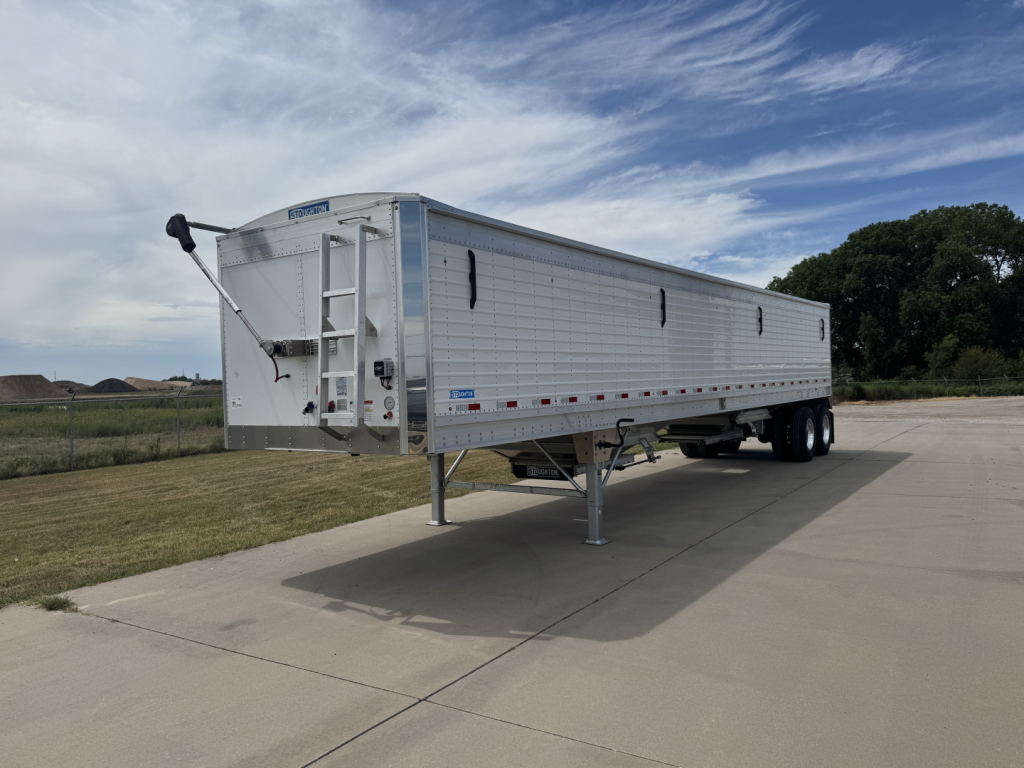 Trailer side view showing 42' aluminum hopper with corrugated sidewalls and front landing gear. VIN tag visible, Stoughton hopper trailer in a parking lot.
