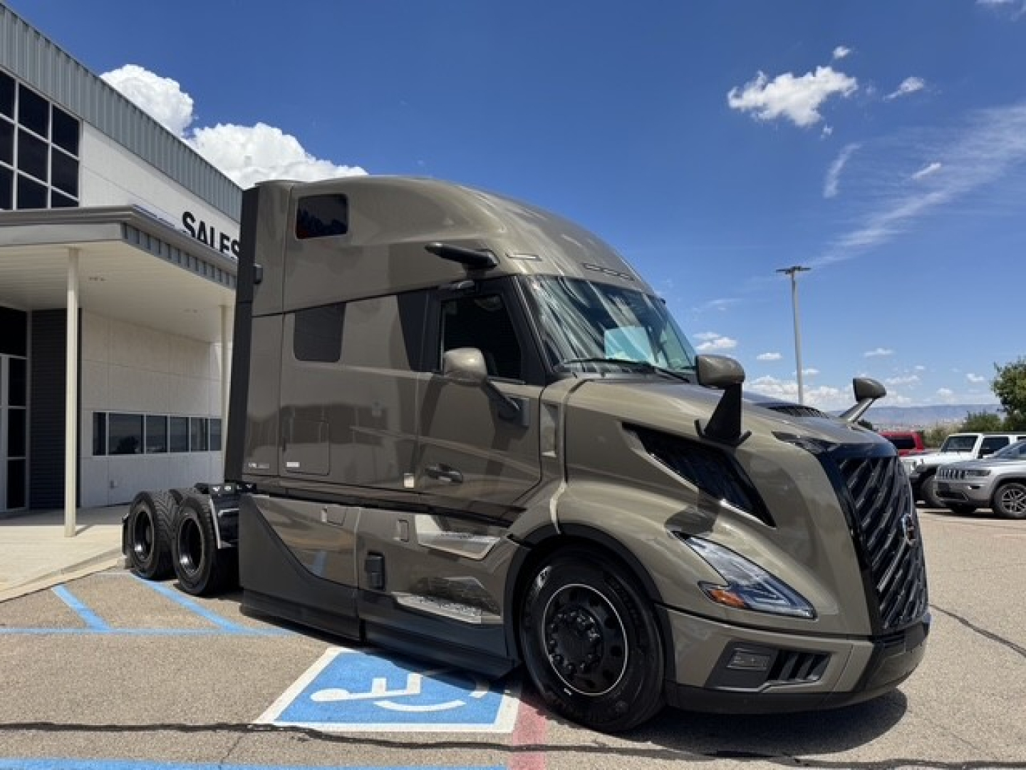 Volvo VNL64T860 truck in a dealership lot, side view with tan brown paint, sleeper cab, three-axle configuration, large front grille.