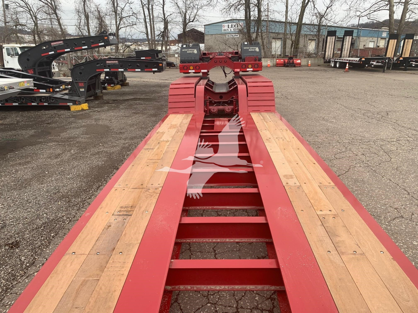 Trailer chassis showing wooden deck and red steel frame, Fontaine 55Lcc lowboy trailer with pin coupling at far end, outdoor lot with other trailers in background.