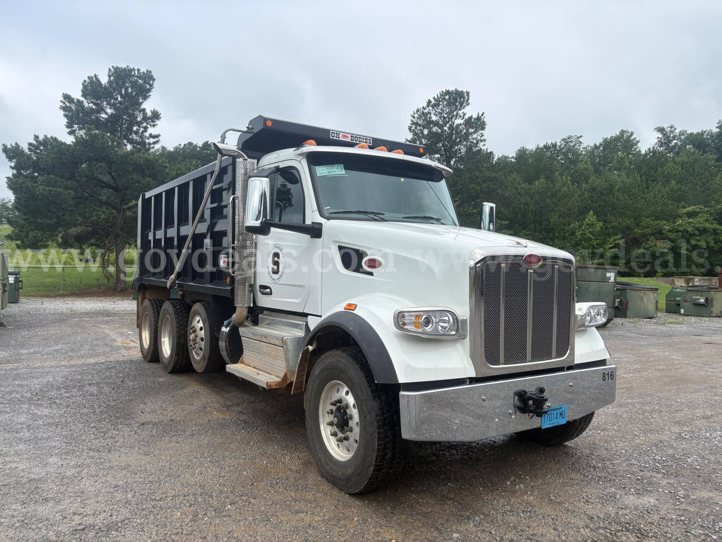 Peterbilt dump truck in a gravel lot, white cab with black dump bed, front grille with Peterbilt badge, multiple axles, chrome bumper, headlights, and side mirrors visible.