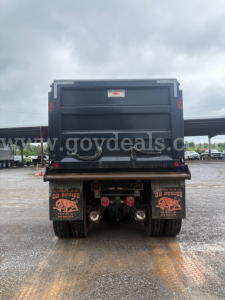 Rear view of a 2025 Peterbilt 377 dump truck showing dump bed and rear axles with dual tires; mud flaps with logos; platform and hitch area visible.