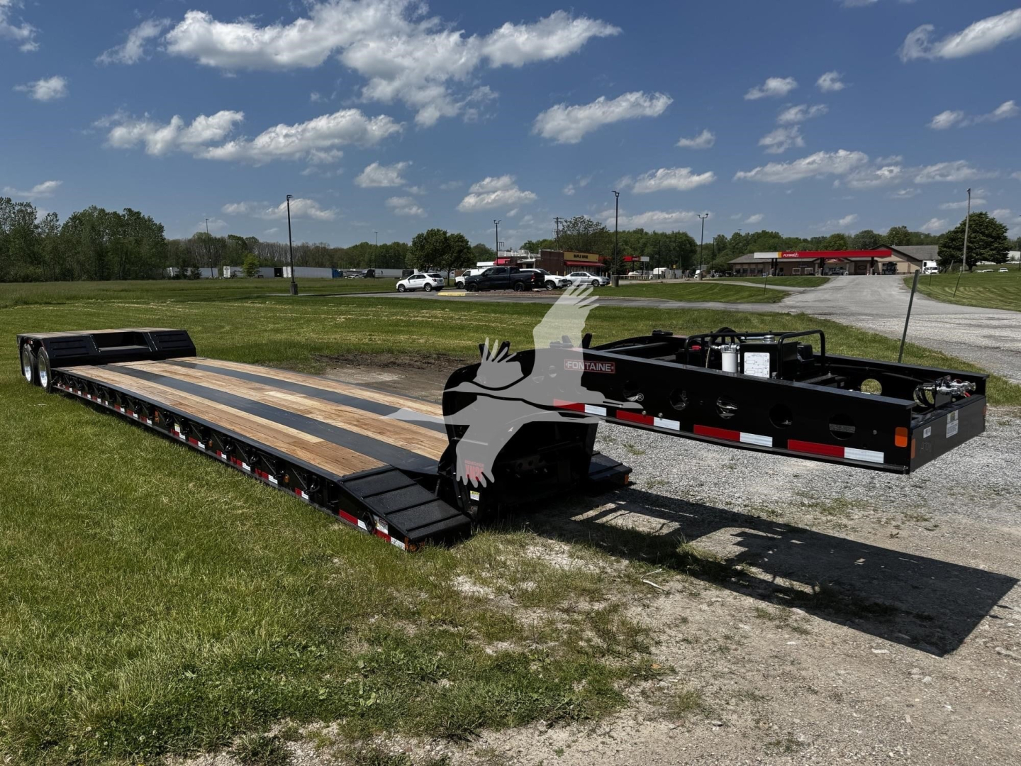 Fontaine Renegade lowboy trailer with long wooden deck and black frame, viewed from side with ramp folded, shows registration and stock lot in background.