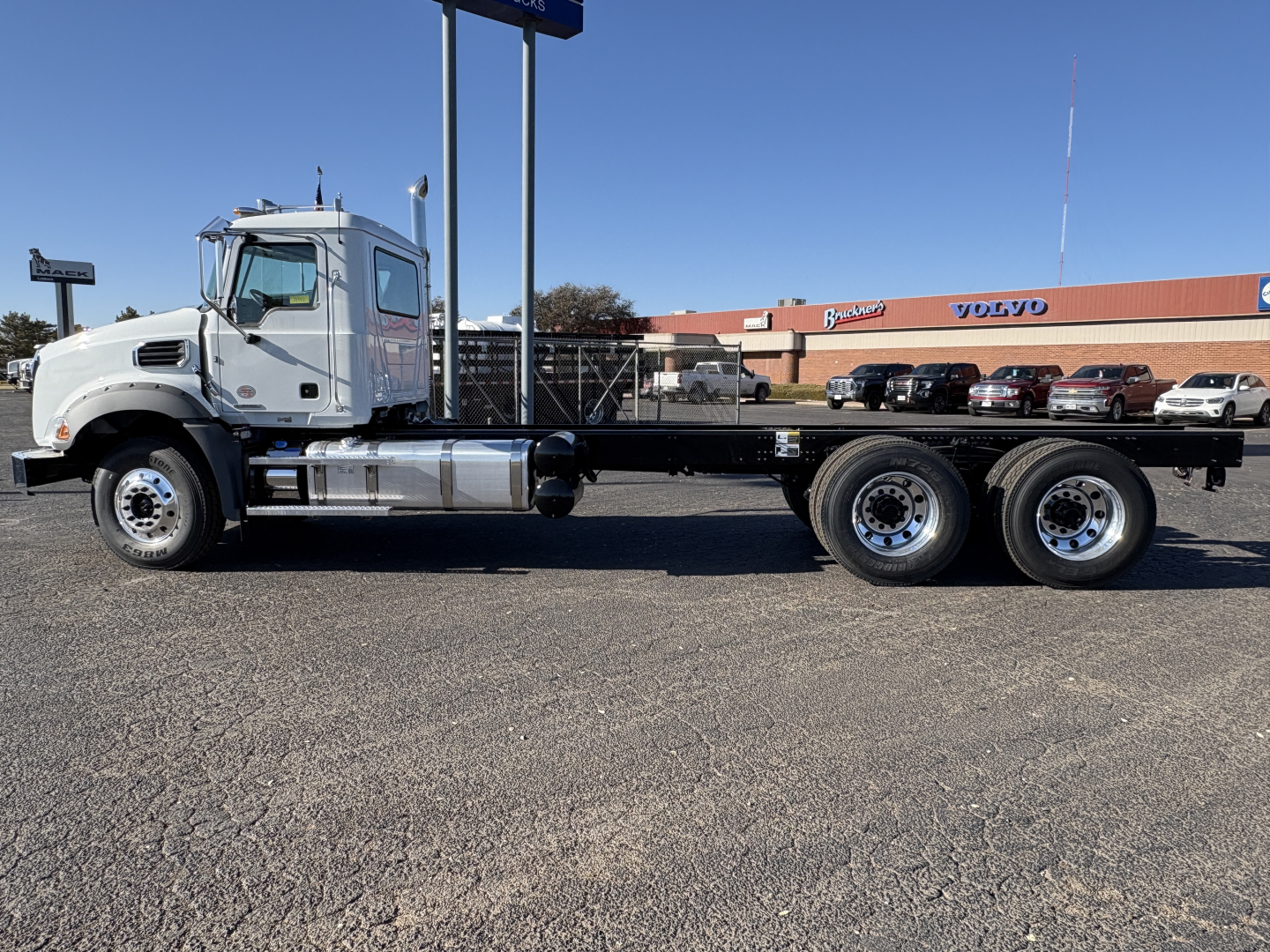 Truck chassis with long wheelbase and three rear axles, flatbed frame, Mack GR64B cab forward truck profile, VIN plate area not visible, WINCH- Oil Field category signage in background.