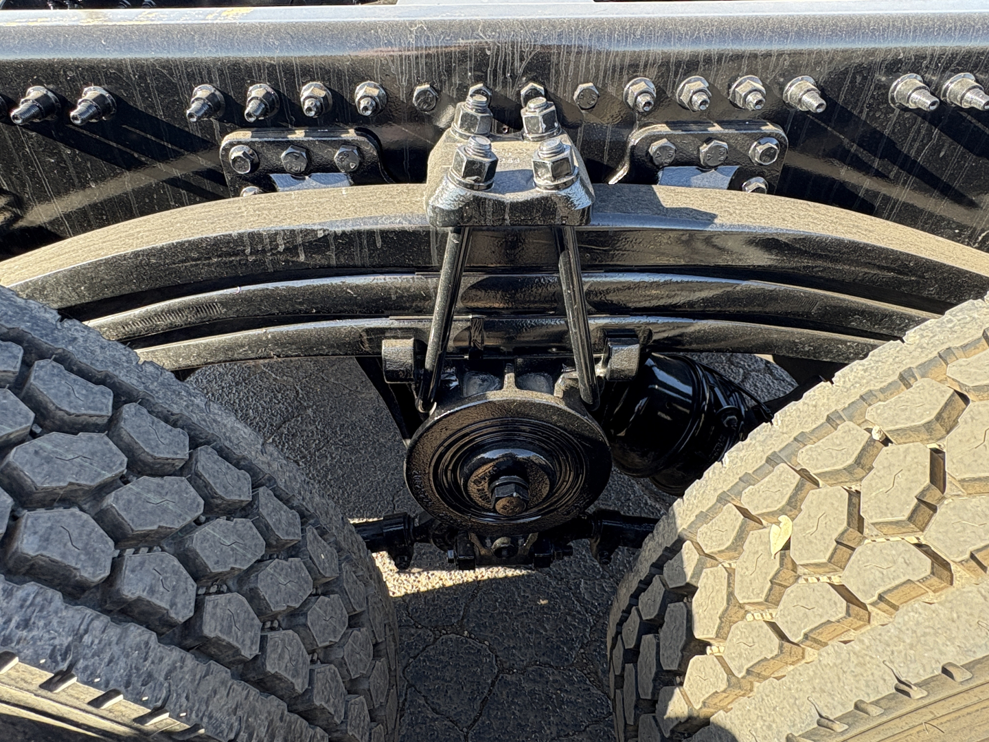 Front view undercarriage of a 2027 Mack GR64B showing suspension components and drive axle, with dual rear wheels and a belt/pulley assembly.