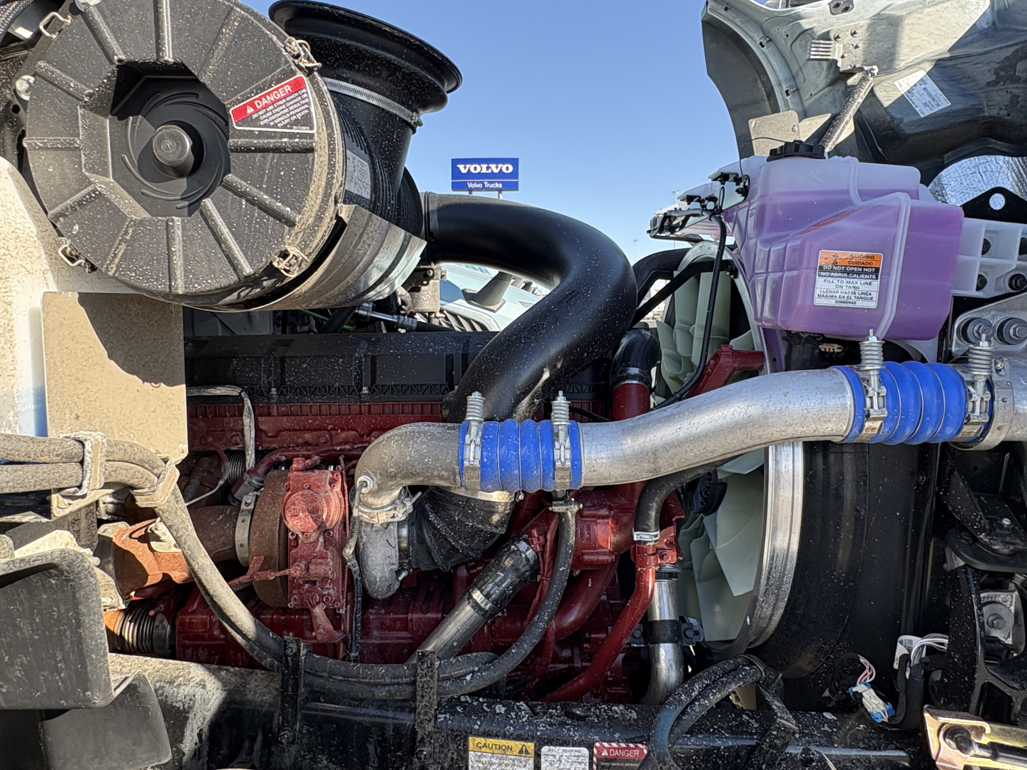 Engine bay of a 2027 Mack GR64B showing red engine block, blue coolant hoses, pink reservoir, and black air intake with warning labels.