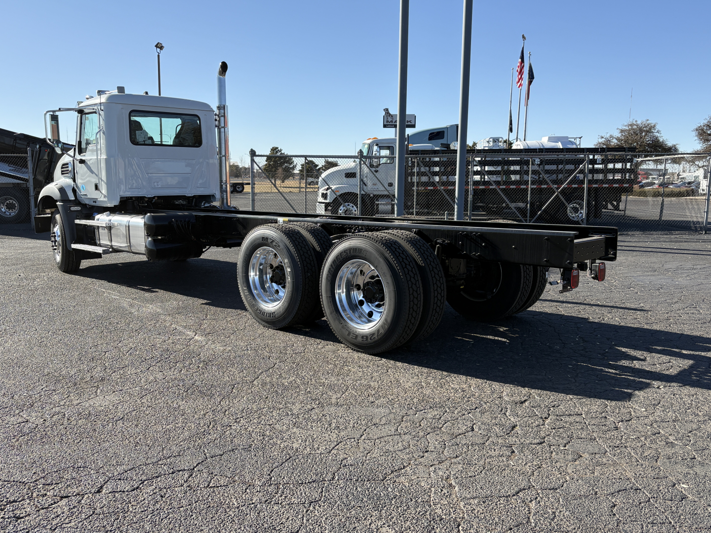 Truck frame with flatbed and dual rear axles in a dealership lot; cab visible left, no load on bed.