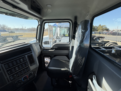 Interior cab view of a 2027 Mack GR64B truck showing steering wheel, dashboard, and cloth seat with plastic cover; window and mirror reflections visible.