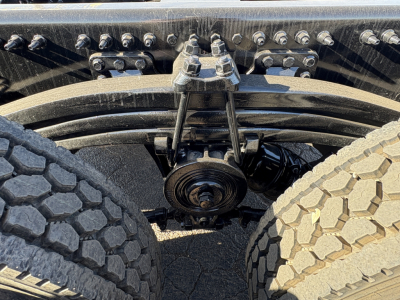 Front view undercarriage of a 2027 Mack GR64B showing suspension components and drive axle, with dual rear wheels and a belt/pulley assembly.