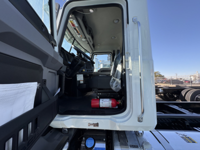 Interior of a new Mack GR64B truck cab showing driver’s seat, steering area, and a red fire extinguisher on the floor; open door and exterior cab visible.