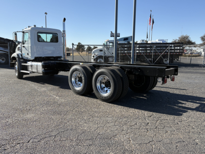 Truck frame with flatbed and dual rear axles in a dealership lot; cab visible left, no load on bed.