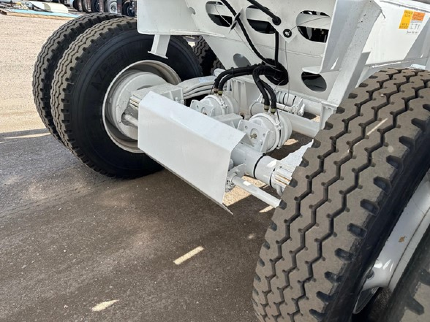 Bottom-dump trailer drivetrain with dual tires and undercarriage components visible; hydraulic lines and linkage near white-painted chassis.