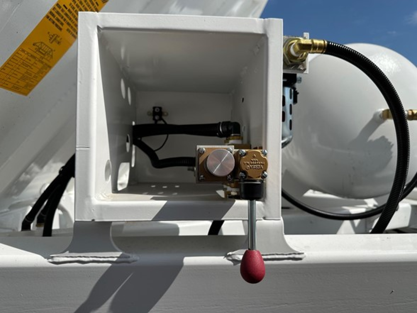 Dump trailer component showing hydraulic control valve with red knob and white square hopper section, side tank and hoses visible.