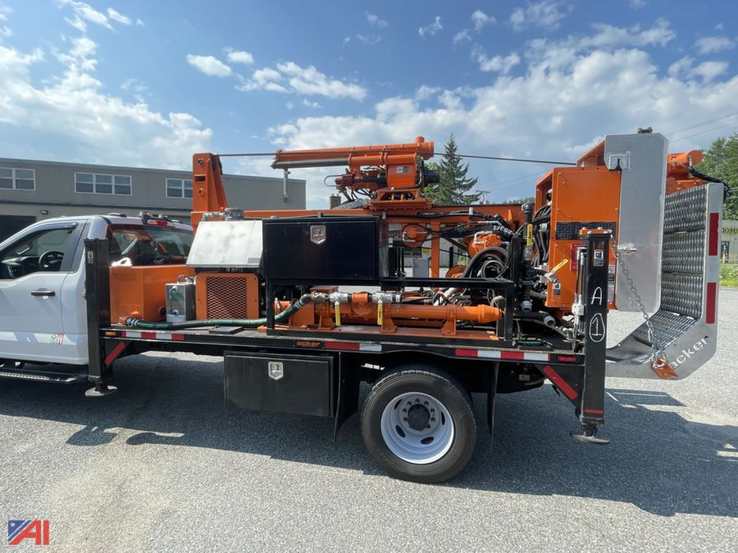Truck-mounted drill rig with orange equipment on a flatbed utility truck showing control panels, hoses, and a drill head.