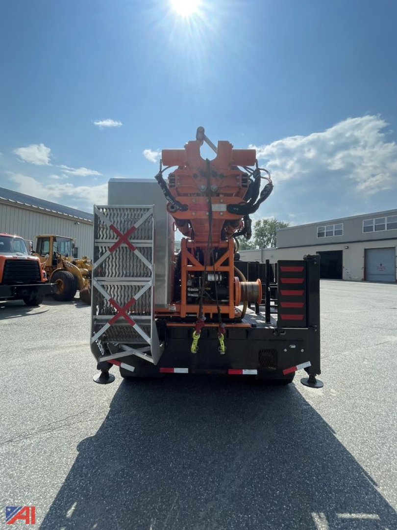 Back view of a mounted drill rig on a Ford F600 utility truck. Bright orange equipment with hoses and controls, metal steps on left, storage box on lower right, clear sky.