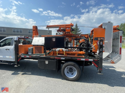 Truck-mounted drill rig with orange equipment on a flatbed utility truck showing control panels, hoses, and a drill head.