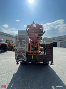 Back view of a mounted drill rig on a Ford F600 utility truck. Bright orange equipment with hoses and controls, metal steps on left, storage box on lower right, clear sky.