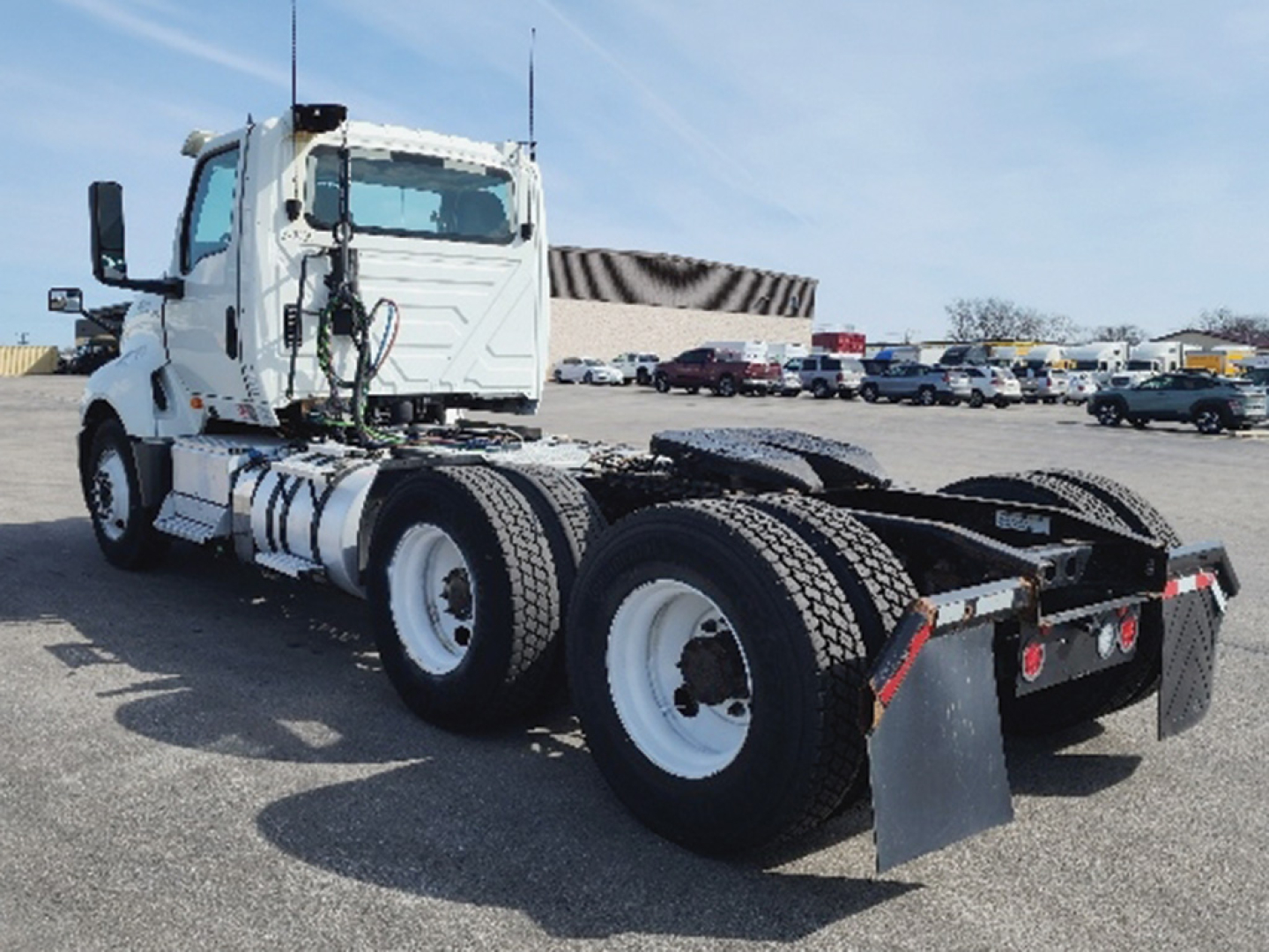 Truck chassis with cab in white and dual rear axles, flatbed rear with mud flaps, showing fifth-wheel hitch area and dual rear tires.