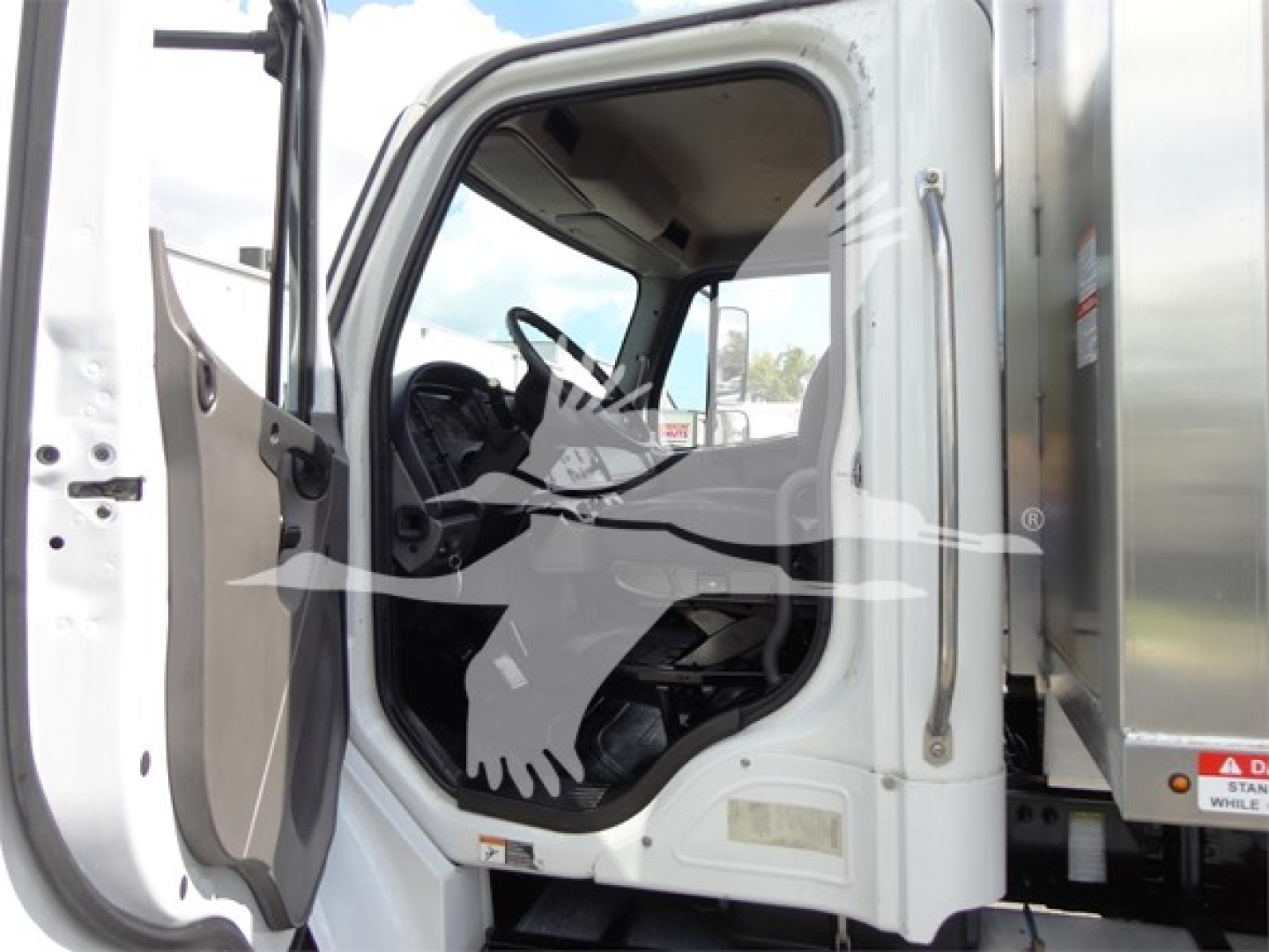 Truck cab interior with open door of a Freightliner M2 106, steering wheel visible, dashboard details, and a metallic dump body adjoining the cab.