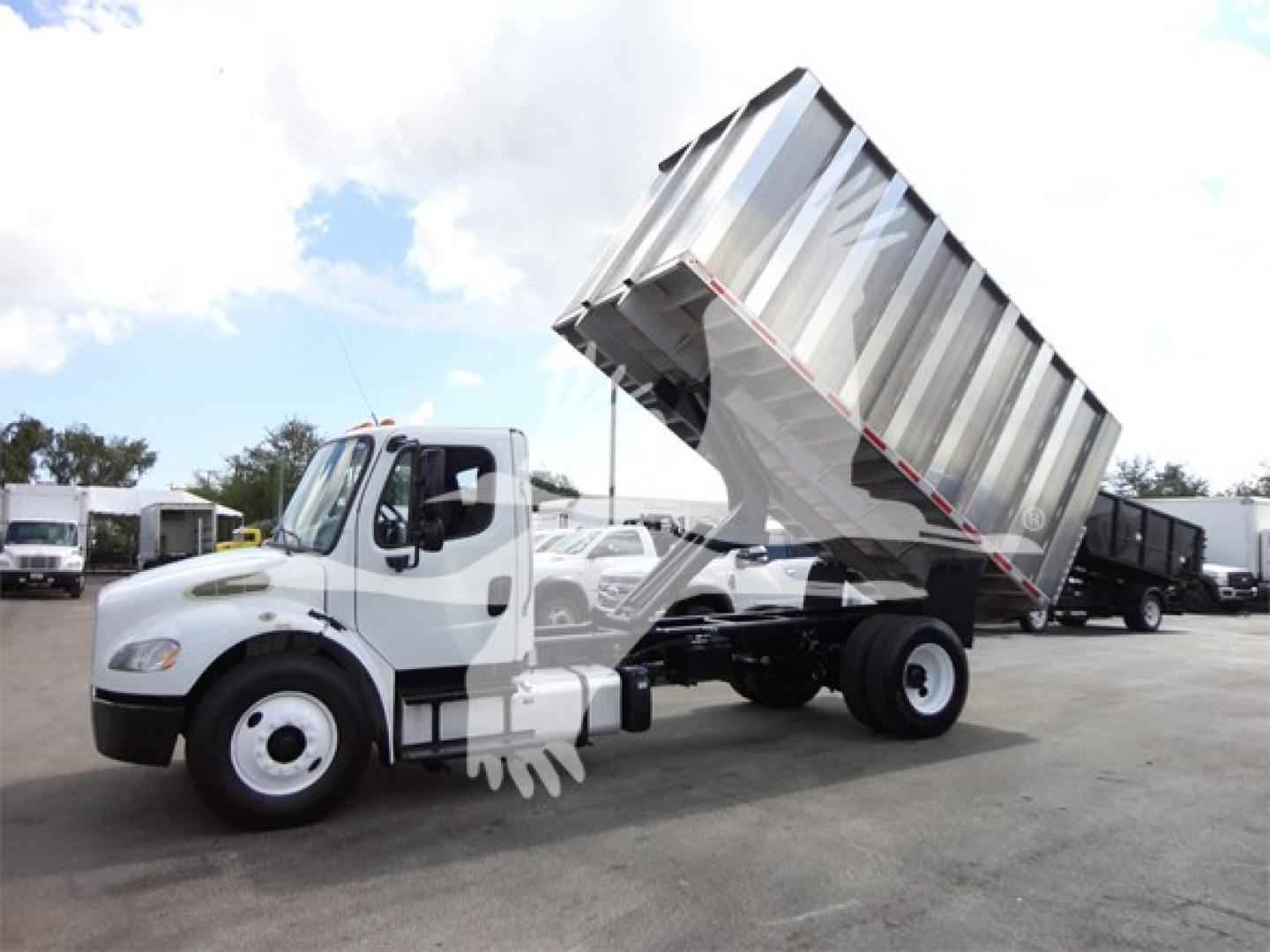 Truck chassis with a tilted aluminum dump bed on a Freightliner M2 106 in a dealership lot.