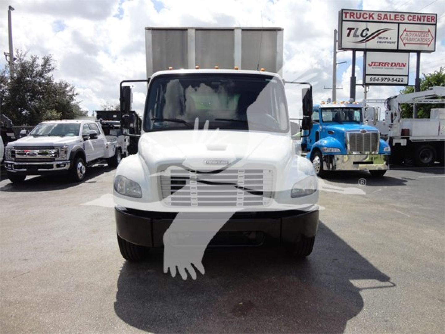 Front view of a white Freightliner M2 106 truck in a lot with other vehicles and a large cargo container behind.
