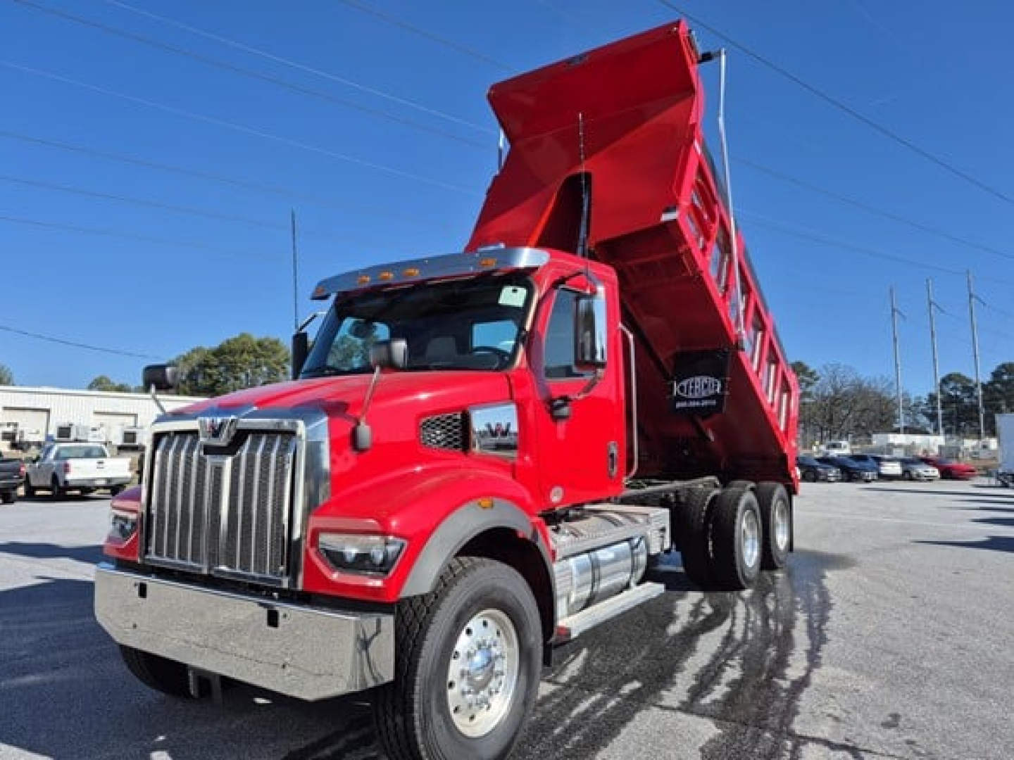 Three-quarter view of a red Western Star dump truck with its bed raised. Visible text on cab may include branding and decals, and the vehicle sits in a lot with other trucks.