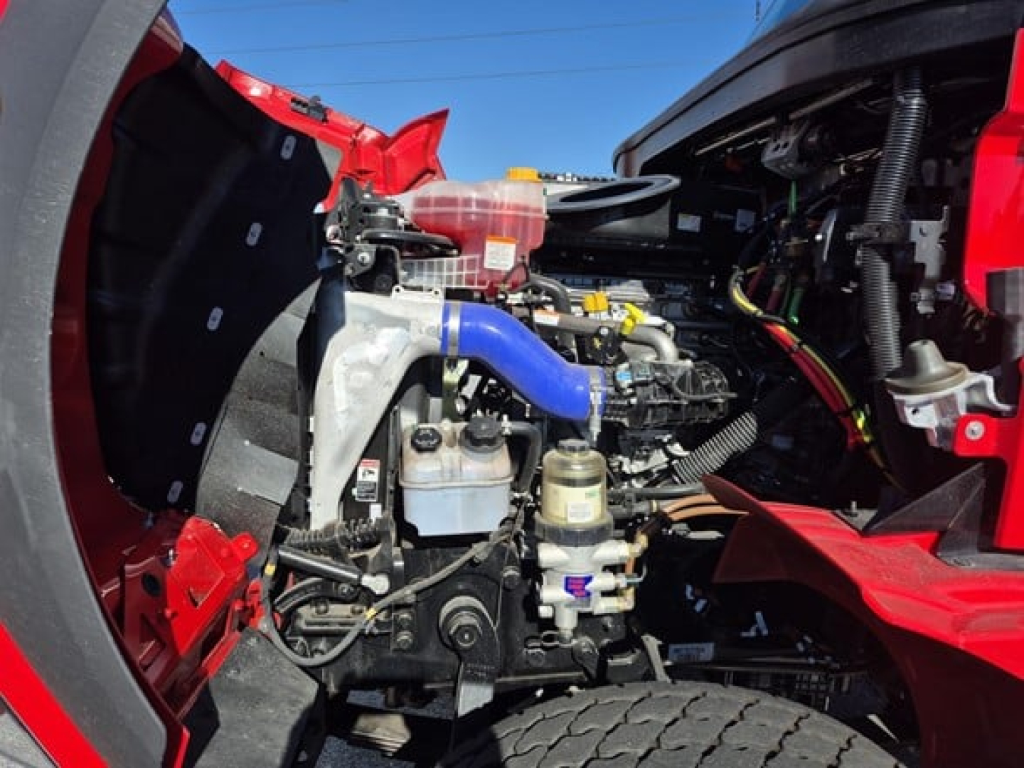 Truck engine bay of a 2026 Western Star 47X showing blue coolant hose, auxiliary reservoir, and various engine components.