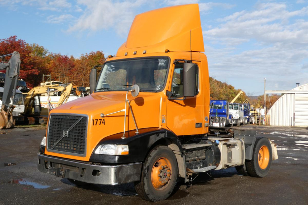 Volvo VNM42T conventional day cab truck in orange, front view with raised sleeper cab, VIN tag and stock number nearby.