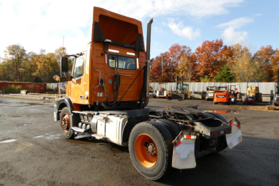 Volvo VNM42T conventional daycab tractor viewed from rear-left, orange paint, visible cab interior, rear fifth wheel, dual rear tires, VIN plate area not legible.