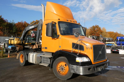 Volvo VNM42T conventional daycab truck with orange cab and hood, front grille, side mirrors, and a yellow excavator in background.
