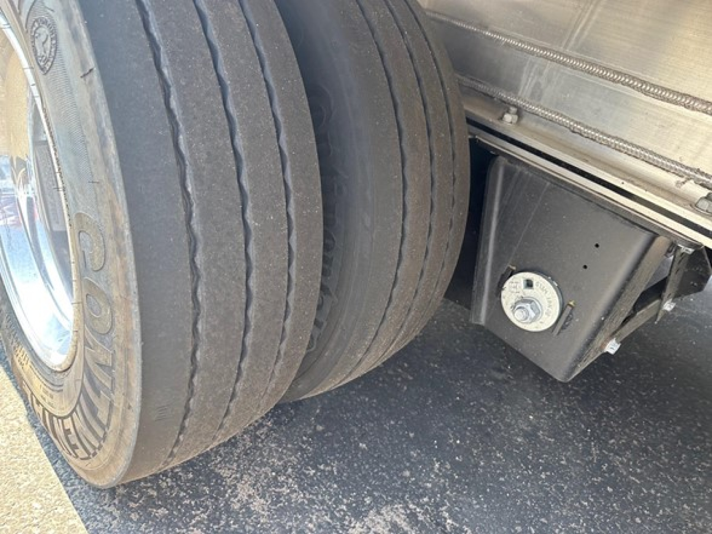 Dual rear tires of a Mac drop deck trailer showing wheel, suspension, and frame with a square hydraulic or air tank/box housing near the axle.