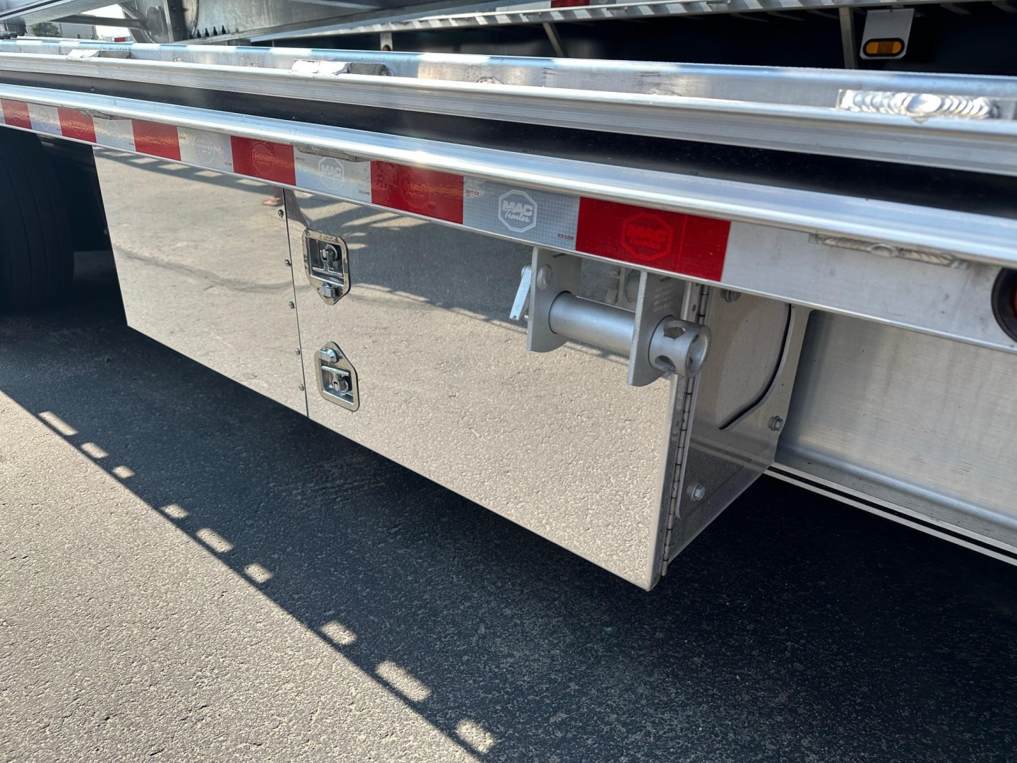 Drop deck trailer side view showing reflective red/white tape, polished aluminum toolbox, and towing hitch with pins.