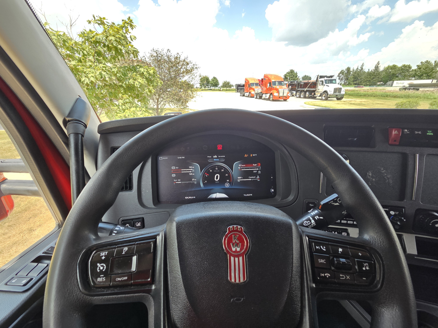 Kenworth T680 cab interior steering wheel with dashboard; visible text on dash and steering controls, outside shows trucks in a yard.