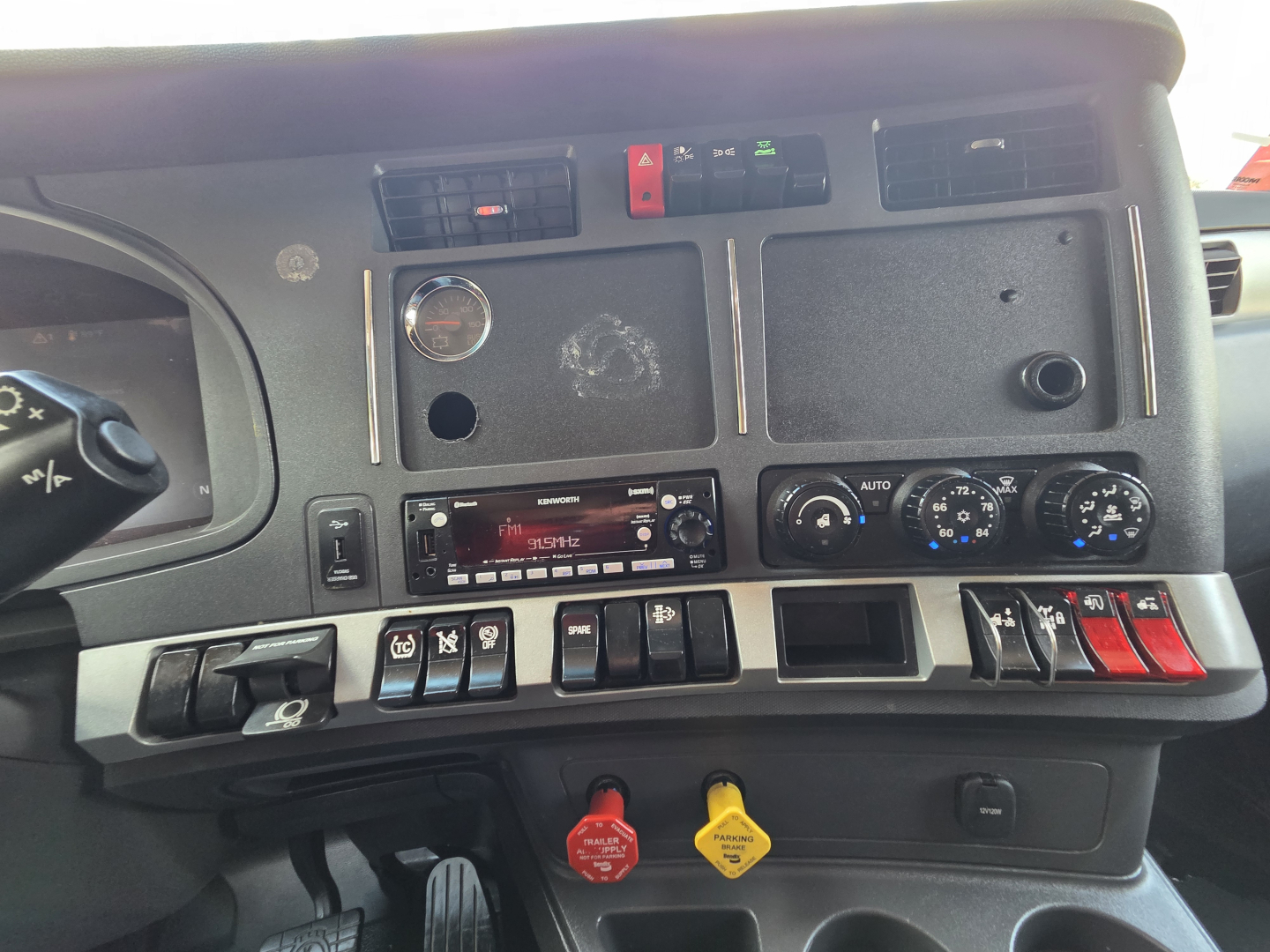 Dashboard of 2023 Kenworth T680 with switches, gauges, and control panels visible; includes red and yellow push buttons, climate controls, and radio unit.