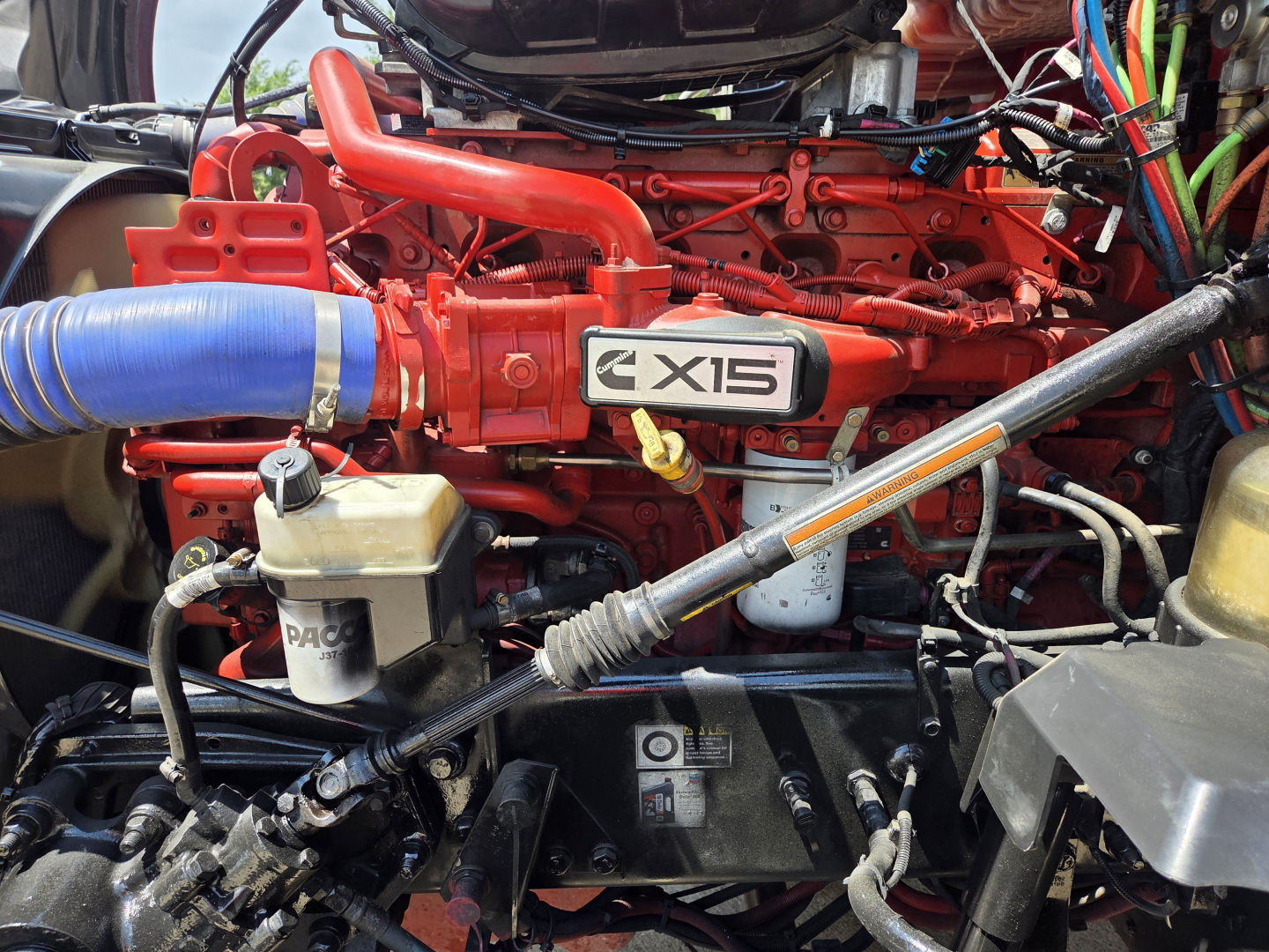 Kenworth T680 engine bay with red X15 badge, blue intercooler hose, and various hydraulic lines.