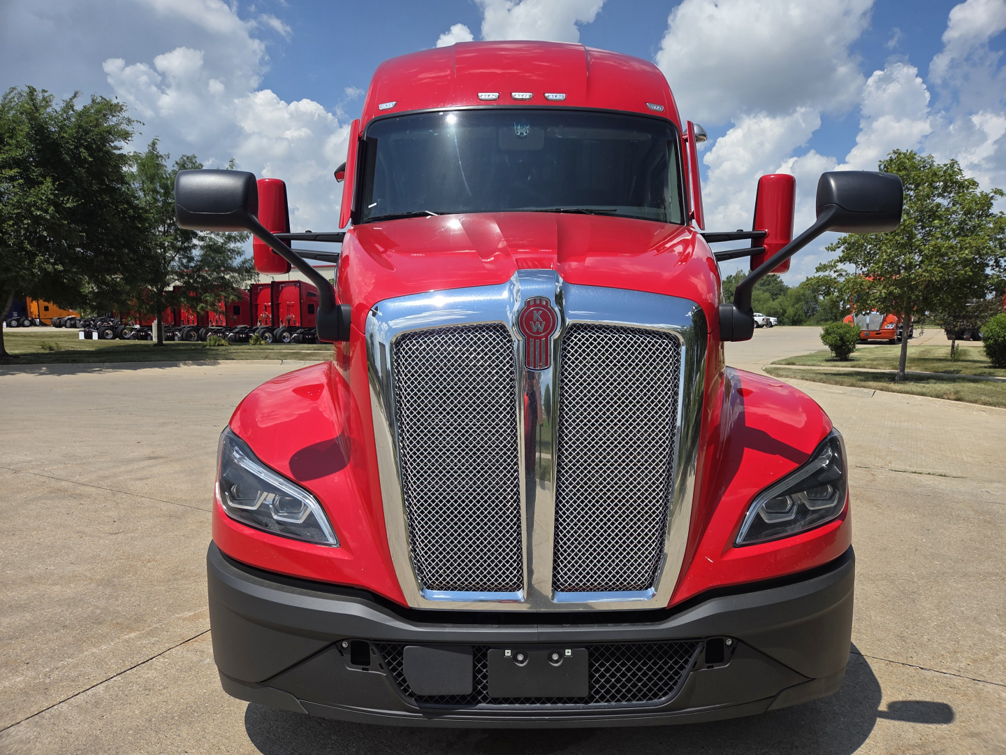 Kenworth T680 front view showing chrome grille, red cab, black bumper, and side mirrors in a parking lot.