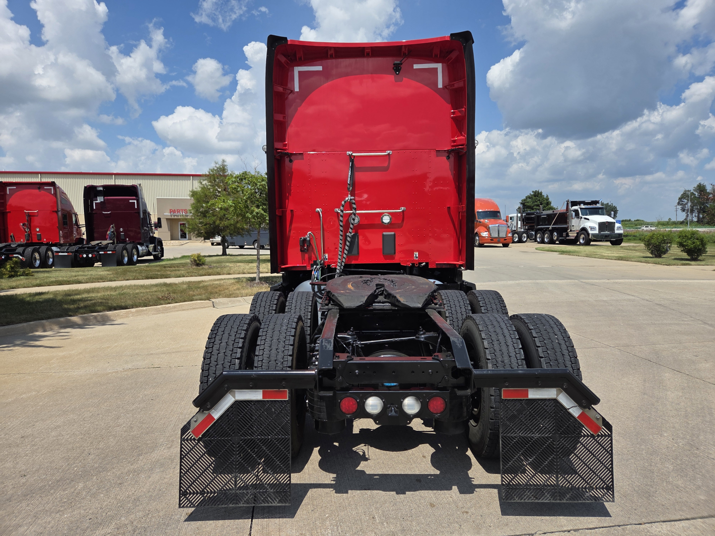 Kenworth T680 rear view showing fifth wheel, dual rear axles, mud flaps, and chassis with red sleeper cab in background.