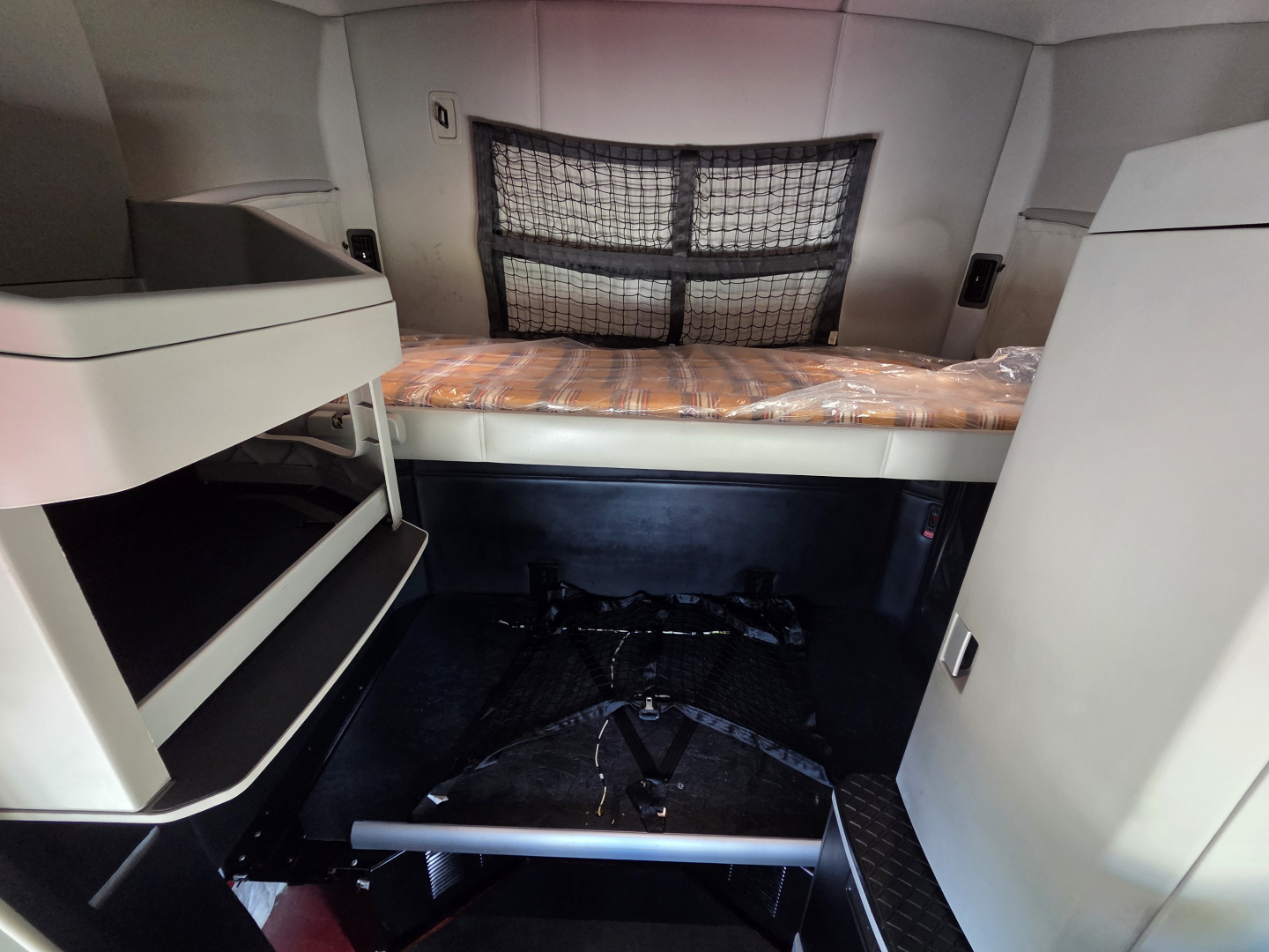 Kenworth T680 sleeper cab interior showing bed with orange plaid bedding, overhead storage shelves, and mesh partition behind; cabin floor with a folded step and interior panels.