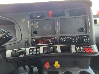 Dashboard of 2023 Kenworth T680 with switches, gauges, and control panels visible; includes red and yellow push buttons, climate controls, and radio unit.