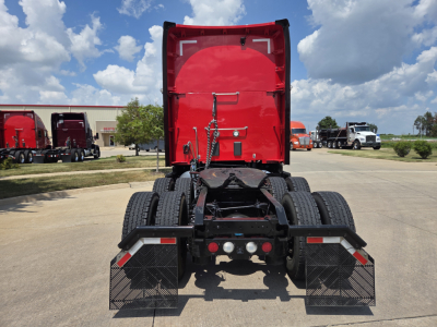 Kenworth T680 rear view showing fifth wheel, dual rear axles, mud flaps, and chassis with red sleeper cab in background.