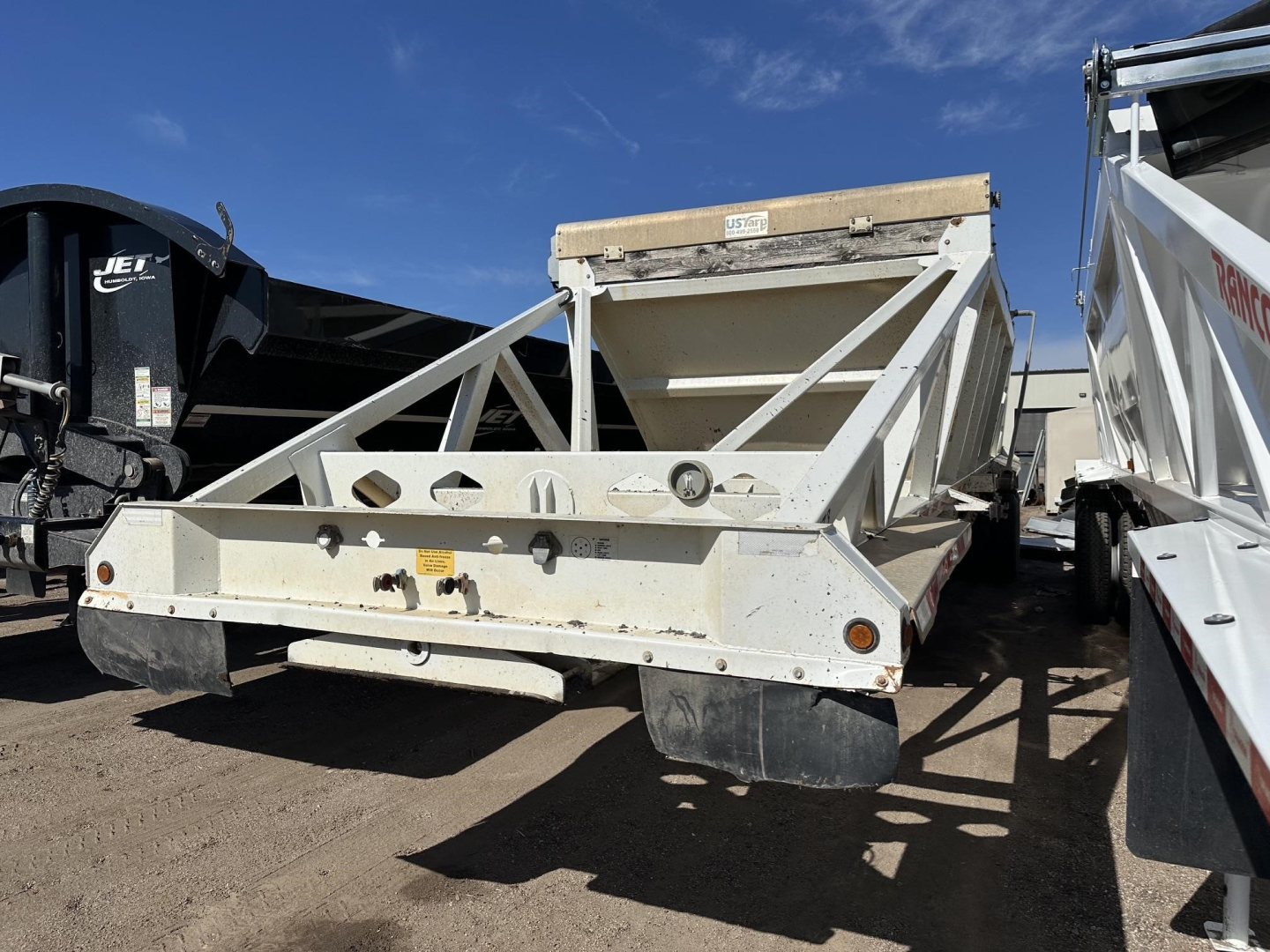 Dump trailer showing steel bottom and electric flip tarp support in a yard; front bumper, side rails, and rear drop area visible.